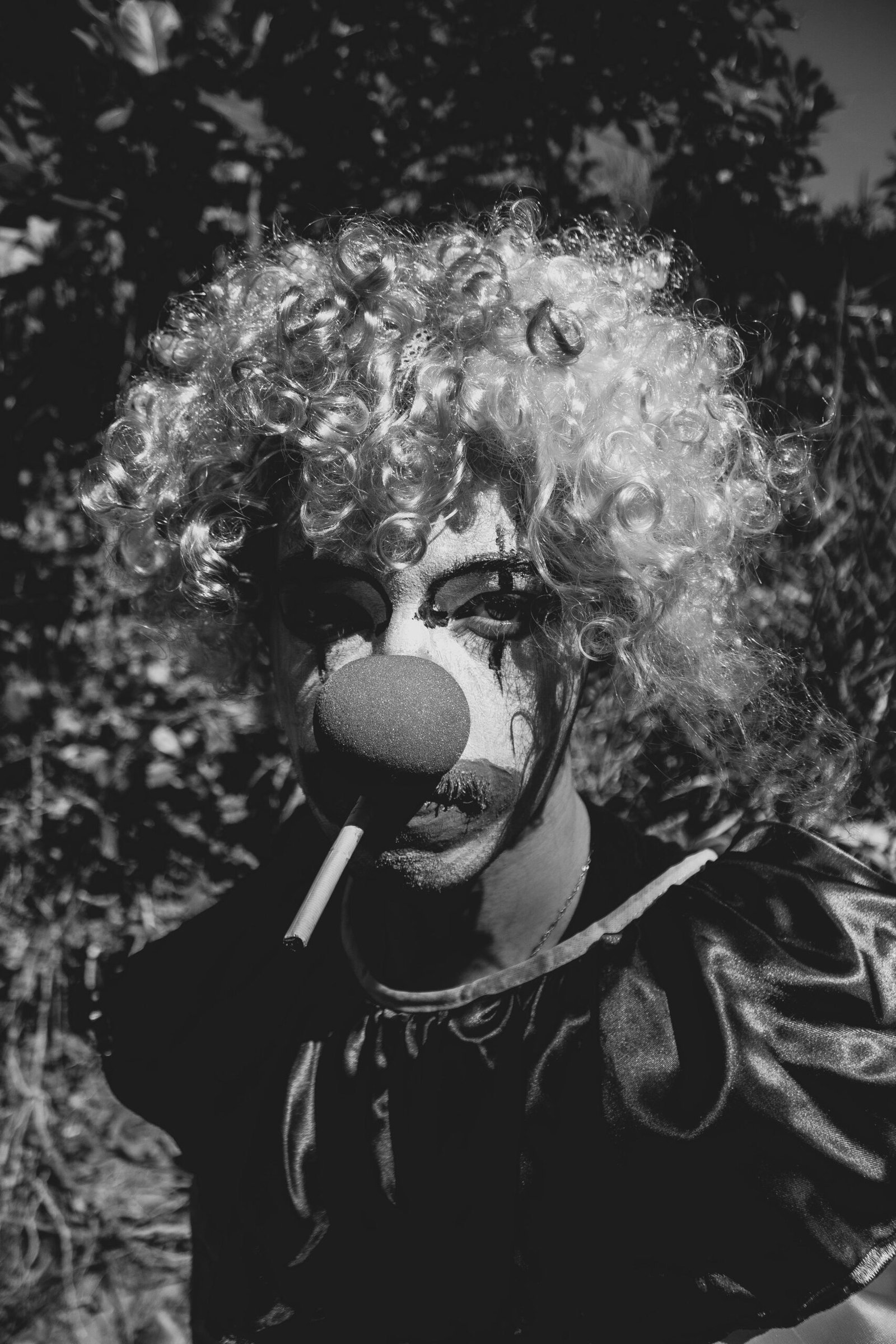 Black and white portrait of a clown with curly hair and face paint, holding a cigarette.